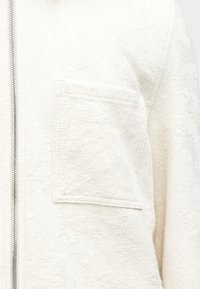 White textured fabric jacket with a square chest pocket and a vertical silver zipper on the left side.