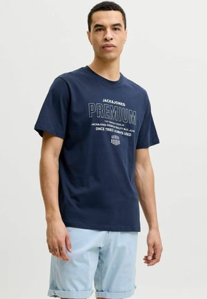 Hombre vistiendo una camiseta gráfica azul marino de Jack & Jones Premium y pantalones cortos azul claro con los bajos remangados, de pie contra un fondo sencillo.