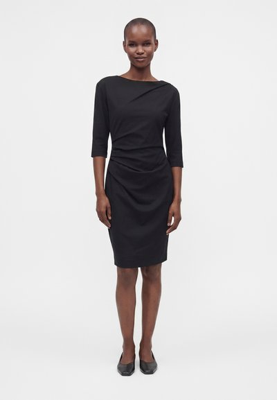 Ψηλή γυναίκα με κοντά μαλλιά που φορά στενό μαύρο φόρεμα μέχρι το γόνατο με μανίκια τριών τετάρτων και μαύρα επίπεδα παπούτσια, στέκεται μπροστά σε απλό φόντο.