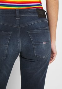 Mörkblå denimjeans har en figursydd design, bakfickor med sömnadsdetaljer och en svart läderlapp med logotyp vid midjan.