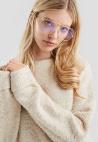 Blondynka w dużych złotych okularach przeciwsłonecznych z niebieskimi szkłami, ubrana w miękki, kremowy sweter z puszystego materiału, trzymająca dłonie blisko klatki piersiowej.