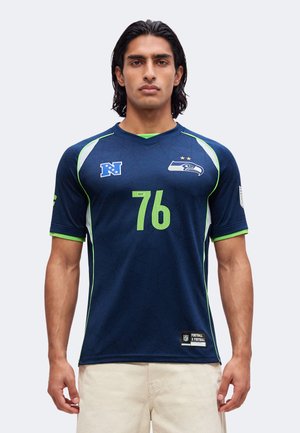 Homme aux cheveux foncés mi-longs portant un maillot bleu marine des Seattle Seahawks avec le numéro 76 et un pantalon beige, debout devant un fond clair uni.