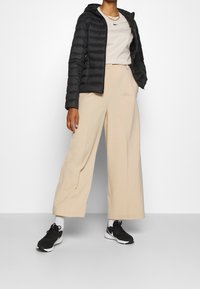 Černá péřová bunda s kapucí, smetanové tričko, béžové kalhoty s širokými nohavicemi a černé sportovní boty s bílými akcenty.