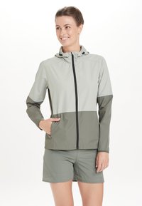 Lichte jas in lichtgrijs en donker olijfgroen, met een capuchon, ritssluiting en zakken. Textiel met textuur en contrasterende kleuren.