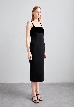 Schwarzes Midi-Kleid mit einer taillierten Silhouette, das ein Samtoberteil, dünne Träger und zwei Seitentaschen aufweist. Kombiniert mit schwarzen Riemchen-Absätzen.