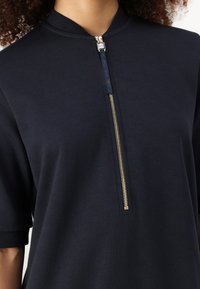 Navy Zip-Up-Top mit hohem Kragen, kurzen Ärmeln und einem goldenen Reißverschluss-Detail an der Vorderseite, aus einem glatten, weichen Material gefertigt.