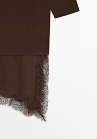 Vêtement en tissu marron avec manches longues et ourlet asymétrique orné de dentelle florale délicate sur fond blanc.