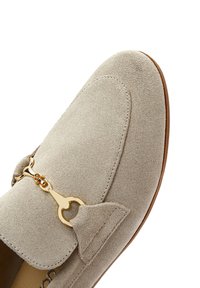 Mocassino in suede di colore beige chiaro, con un accento di hardware dorato, dettagli cuciti e una finitura morbida e testurizzata.