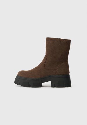 Botas de tornozelo em camurça castanha com um acabamento texturizado, bico arredondado e uma sola de borracha preta grossa com um padrão de tração.