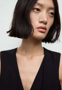Zwarte, mouwloze kleding met een diepe V-halsontwerp. Gladde stofstructuur, aansluitende lijfje en minimalistische stijl. Donker haar, neutrale make-up.