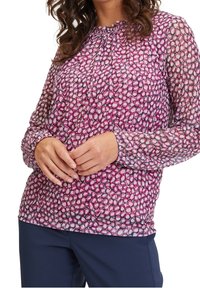 Transparante blouse met lange mouwen in roze met cirkelpatronen. Heeft een geplooide halslijn en elastische manchetten. Gecombineerd met marineblauwe broek.