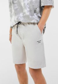 Shorts beige clair avec une taille élastique et un cordon de serrage noir. Dotés de poches latérales et d'un petit détail imprimé noir sur la jambe gauche.