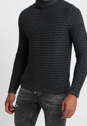 Uomo che indossa un maglione a collo alto grigio scuro con trama e jeans neri scoloriti, in piedi con le braccia rilassate ai lati.