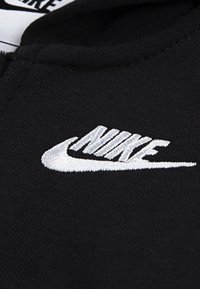 Črn material z belim vezenim Nike logotipom. Tekstura je gladka in mehka, z jasnimi šivi in detajli.