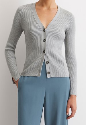 Kobieta w dopasowanym, jasnoszarym, prążkowanym kardiganie z czarnymi guzikami i luźnych niebieskich spodniach, stojąca z ręką w kieszeni.