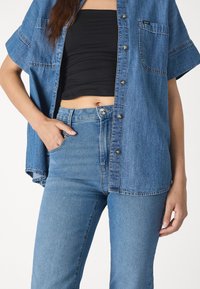 Camisa de mezclilla, de botones y de manga corta, en azul claro, combinada con jeans azul de tiro alto. Textura suave, detalle de bolsillo en la parte frontal.