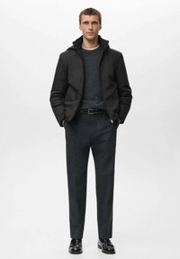 Giacca nera con collo a listino, maglione grigio sotto, pantaloni grigi su misura e scarpe nere, esposti su uno sfondo neutro.