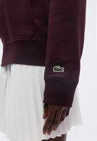 Poignet de sweat-shirt à manches longues bordeaux avec logo crocodile vert au-dessus de la main, associé à une jupe plissée blanche sur un modèle à la peau foncée.