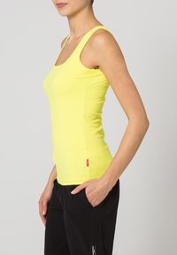 Gul ribbad linne med breda axelband, figurnära design och en liten röd logotypdetalj vid fållen, kombinerat med svarta byxor.