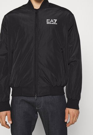 Chaqueta bomber negra con cremallera y logo EA7 Emporio Armani en el pecho izquierdo, usada sobre vaqueros oscuros.