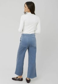 Pantalons en velours côtelé bleu à jambe large avec des côtes verticales, dotés de deux poches arrière. Portés avec un haut ajusté à manches longues blanc et des chaussures foncées.