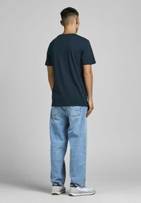 Marineblaues T-Shirt mit kurzen Ärmeln, kombiniert mit hellblauen, lockeren Jeans und grauen Sneakers, von hinten vor einem schlichten Hintergrund betrachtet.
