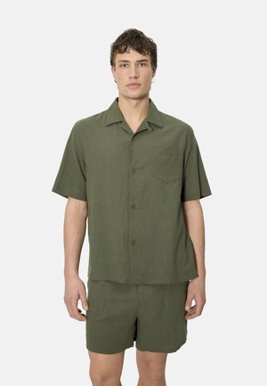 Joven con camisa de manga corta de color verde oliva abotonada y pantalones cortos a juego, de pie frente a un fondo claro liso.