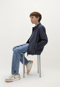 Marineblauwe lichte jas met een staande kraag, gecombineerd met blauwe jeans en beige sneakers met een blauwe accent en logo. Zittend op een metalen kruk.