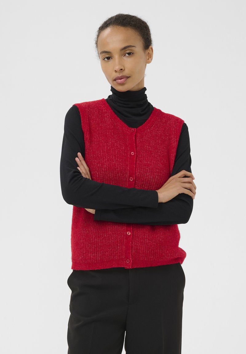 Punainen neulottu liivi, jossa v-kaula-aukko ja nappikiinnitys, teksturoitu pinta, käytetty mustan pitkähihaisen paidan päällä, yhdistetty mustiin housuihin.