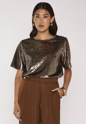 Donna che indossa un top bronzeo con paillettes e maniche corte, pantaloni marroni a vita alta e gamba larga, orecchini a cerchio dorati e braccialetti dorati, con una mano in tasca.