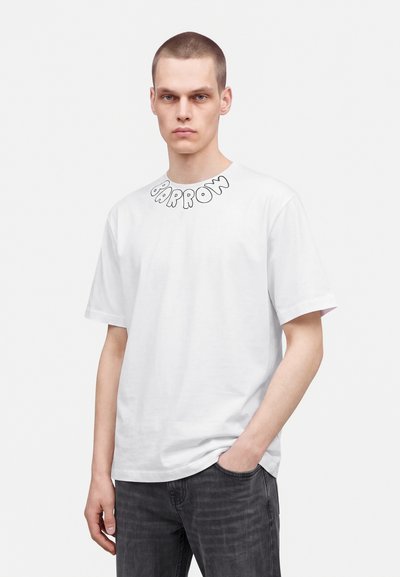 Jeune homme aux cheveux courts portant un T-shirt blanc avec le texte "BARRON" près du col et un jean foncé, debout avec une main dans la poche.
