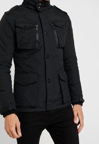 Man som bär en svart jacka med fyra framfickor, två med dragkedjor och två med knappslå, i kombination med svarta byxor.