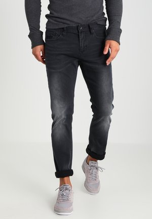 Mann trägt hochgekrempelte schwarze Jeans, dunkelgraues Langarmshirt und hellgraue Reebok-Sneaker, geht vor einfarbigem Hintergrund.