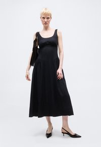 Γυναίκα με κοντά ξανθά μαλλιά που φοράει μαύρο μίντι φόρεμα χωρίς μανίκια, μαύρες γόβες με λουράκι στον αστράγαλο και κρατά μαύρη τσάντα ώμου.