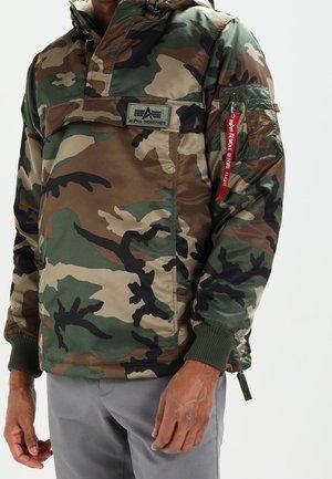 Mężczyzna ubrany w zielono-brązową kurtkę maskującą Alpha Industries z kieszenią na zamek i czerwoną zawieszką „Remove Before Flight” na rękawie.