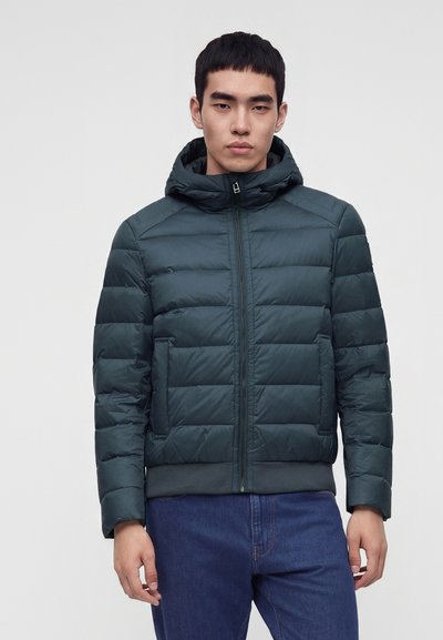 Homme portant une veste matelassée à capuche vert foncé avec fermeture éclair frontale et poches, associée à un jean bleu, debout devant un fond uni.