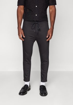 Mężczyzna ubrany w czarne, wąskie spodnie z elastycznym paskiem, białe skarpetki, czarne loafersy oraz czarną, krótko rękawową koszulę z wzorem.