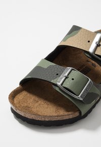 Sandal in tessuto camouflage con due cinturini regolabili, realizzato in pelle testurizzata. Presenta una soletta in sughero e una suola in gomma. Fibbia in metallo color argento.