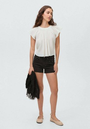Blusa bianca con maniche a sbuffo, abbinata a pantaloni corti di jeans neri. Il modello tiene una sciarpa intrecciata nera e indossa delle ballerine beige. Sfondo neutro.