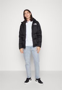 Casaco acolchoado preto com capuz, apresentando um fecho frontal, grandes bolsos e um logotipo no lado esquerdo do peito, vestindo sobre uma camisa cinza clara e jeans.