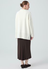 Hvid oversized sweater, brun midi-nederdel med slids bagpå og sorte sko. Glatte teksturer; afslappet, løstsiddende design.