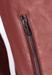 Brunt læder nærbillede med en sølvlynlås og trækstang, der har en tekstureret overflade og kontrasterende syninger langs sømmen.