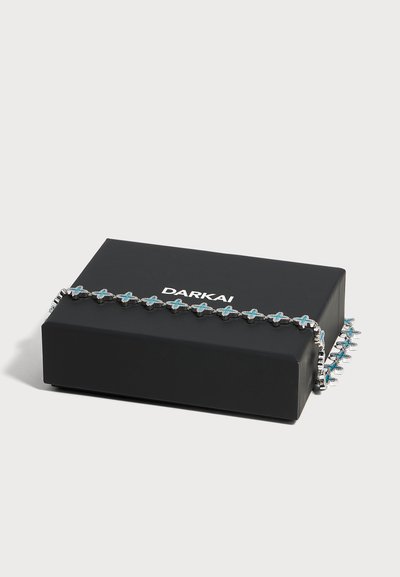 Collier chaîne en argent avec des pierres bleues en forme de croix présenté sur une boîte à bijoux noire DARKAI sur fond blanc.