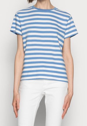 Kobieta ubrana w niebiesko-biały, poziomo paskowany T-shirt z krótkim rękawem oraz białe spodnie, stojąca na jednolitym tle.