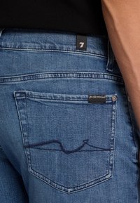 Blaue Denim-Jeans mit einer Gesäßtasche, die ein wellenartiges Stickdesign aufweist. Der Stoff hat eine leicht strukturierte Oberfläche.