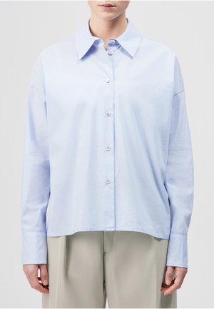 Person mit hellblauem Hemd mit langen Ärmeln und beiger Hose, stehend vor einem schlichten Hintergrund.