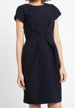 Frau trägt ein figurbetontes schwarzes knielanges Kleid mit kurzen Ärmeln und plissierten Details in der Taille.