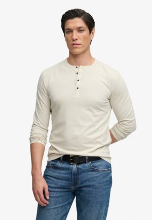 Joven con cabello oscuro que lleva una camisa henley beige de manga larga y vaqueros azules con un cinturón negro, de pie frente a un fondo blanco.