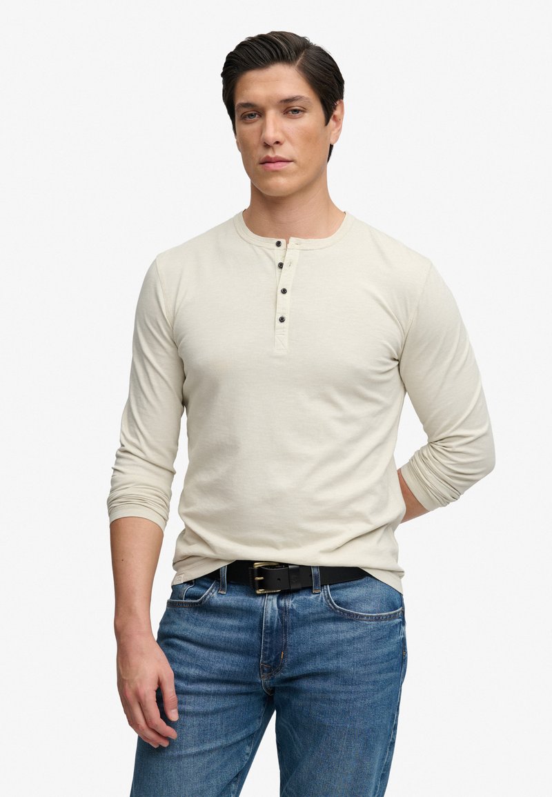Jonge man met donker haar, gekleed in een beige henley met lange mouwen en blauwe spijkerbroek met een zwarte riem, staand tegen een witte achtergrond.