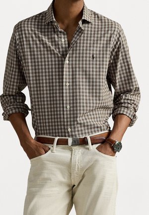 Hombre con camisa marrón de cuadros abotonada con las mangas remangadas, pantalones beige, cinturón marrón y reloj de pulsera, con las manos en los bolsillos.